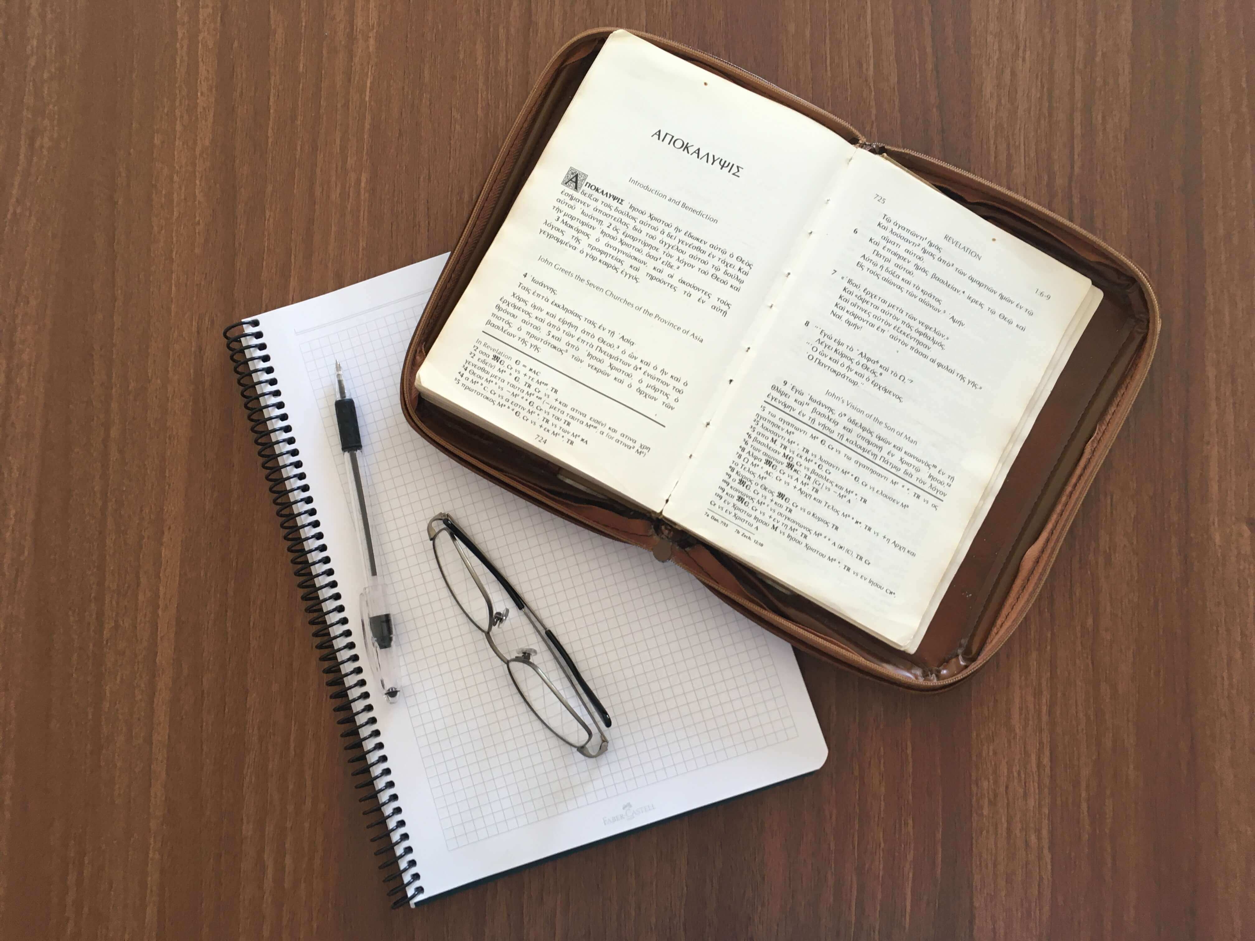 a notebook, a pen, a pair of glasses, and a Greek New Testament opened to Revelation 1